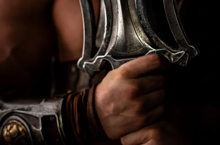 A muscular man holds a large, metal sword in a dark setting.