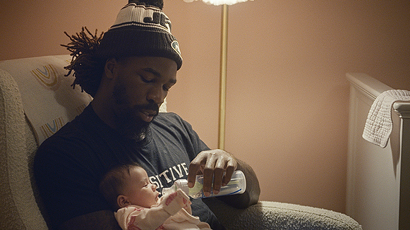 A man feeds his infant child while wearing a NY Jets beanie