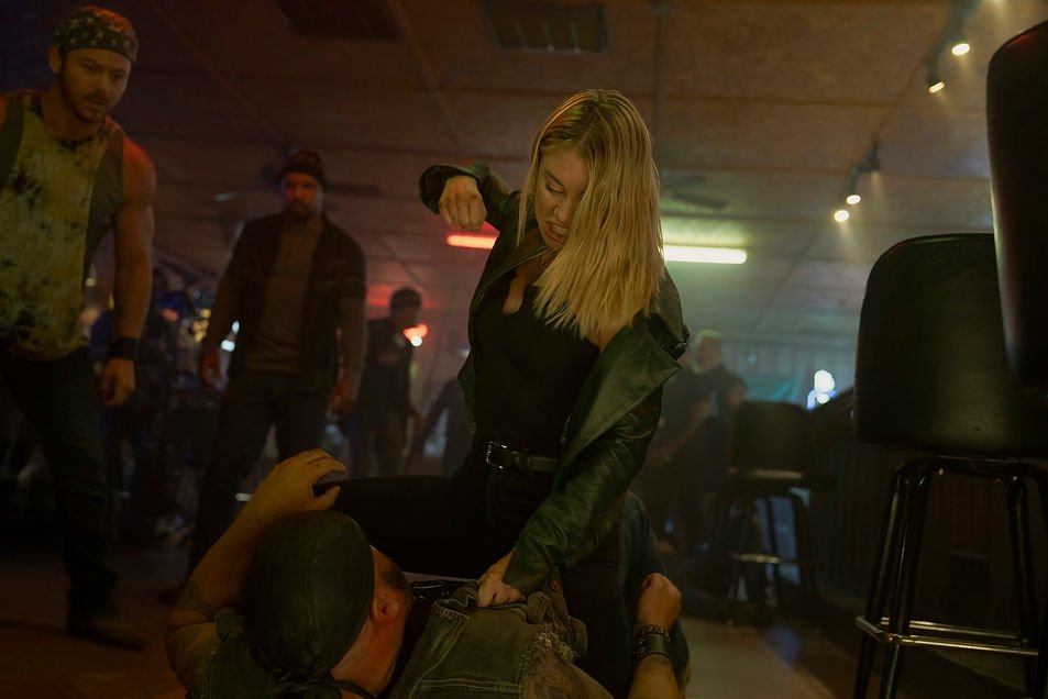 Jennifer Holland as Emilia Harcourt punching a man on the floor in a bar.