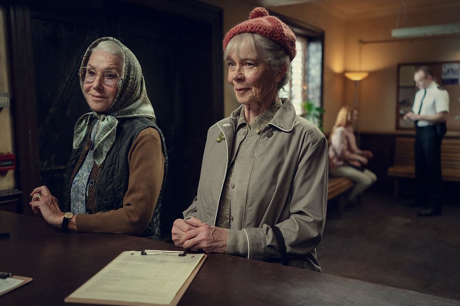 Helen Mirren as Elizabeth in a disguise with Celia Imrie as Joyce standing at a desk with smirks on their face