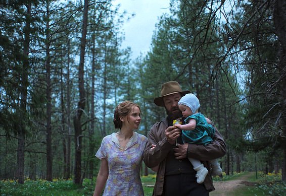 Felicity Jones as Gladys and Joel Edgerton as Robert Grainier with their daughter Kate, stood within a forest, in Train Dreams.