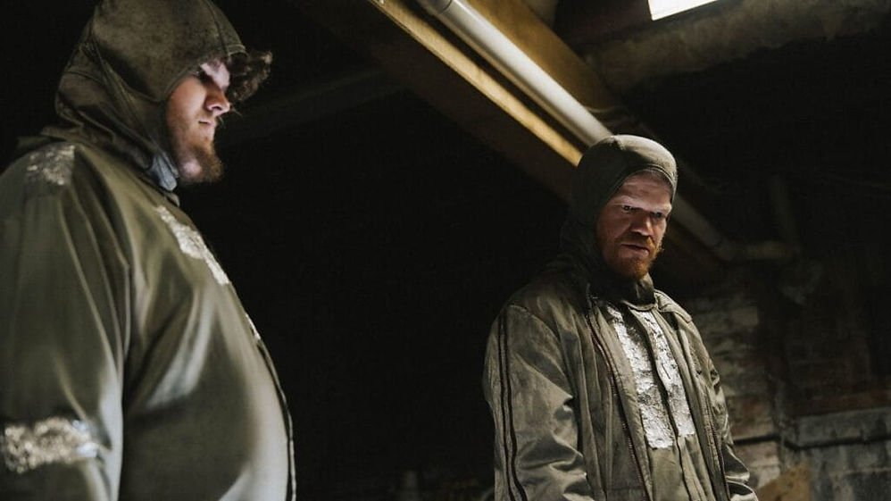 Aidan Delbis as Don and Jesse Plemons as Teddy wearing protective suits while staring at something offscreen in 'Bugonia'