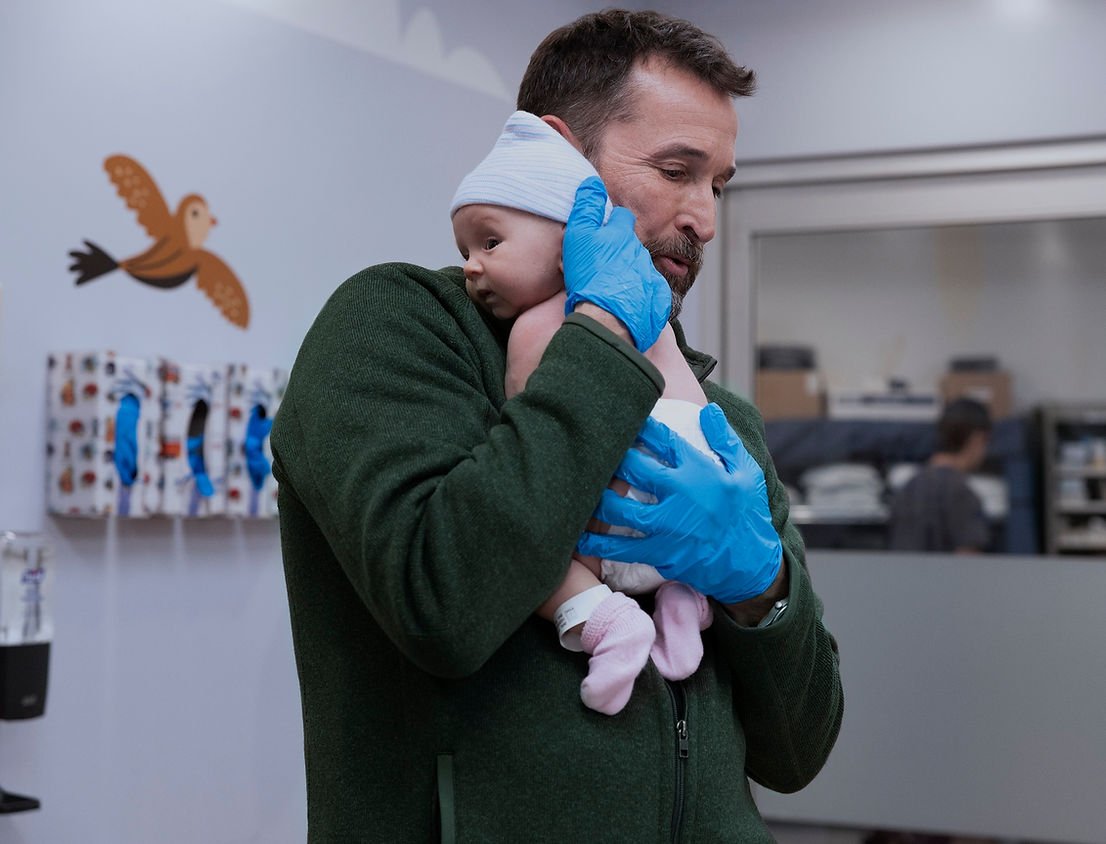 Noah Wyle as Dr Michael "Robby Robinavitch holding a baby in The Pitt Season 2.