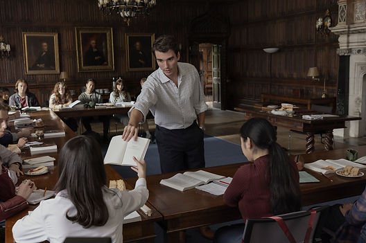 Corey Mylchreest in 'My Oxford Year' handing Sofia Carson a book in a classroom.