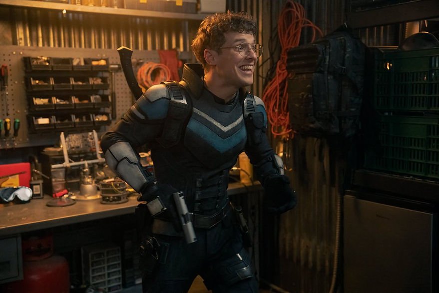 Freddie Stroma as Vigilante 2 in his suit holding a gun and smiling in 'Peacemaker' Season 2