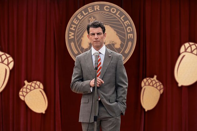 Max Greenfield as Berenger stood on stage in front of a Wheeler College sign in front of a microphone in 'A Man on the Inside' Season 2.