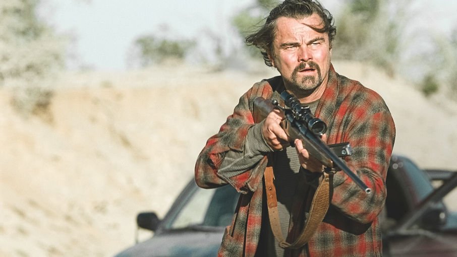 Leonardo DiCaprio as Bob Ferguson standing with a sniper rifle in his robe staring at someone offscreen in 'One Battle After Another'.
