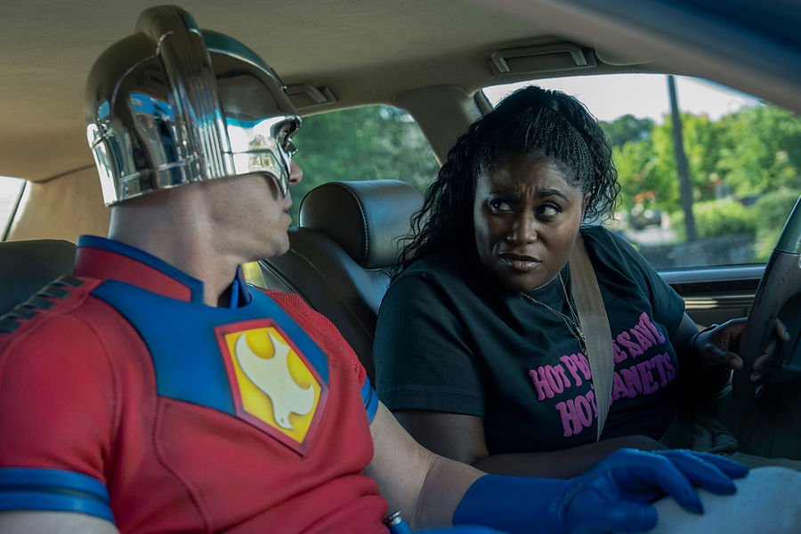 Danielle Brooks as Adebayo siting in her car talking to John Cena as Peacemaker wearing his full suit also sitting in the car