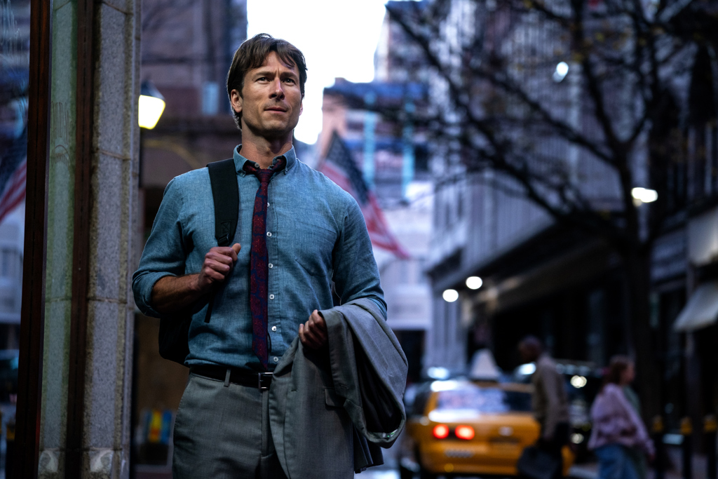 Glenn Powell as Beckett standing on a city street with a backpack over one shoulder and a jacket draped over his other arm in a scene from How to Make a Killing