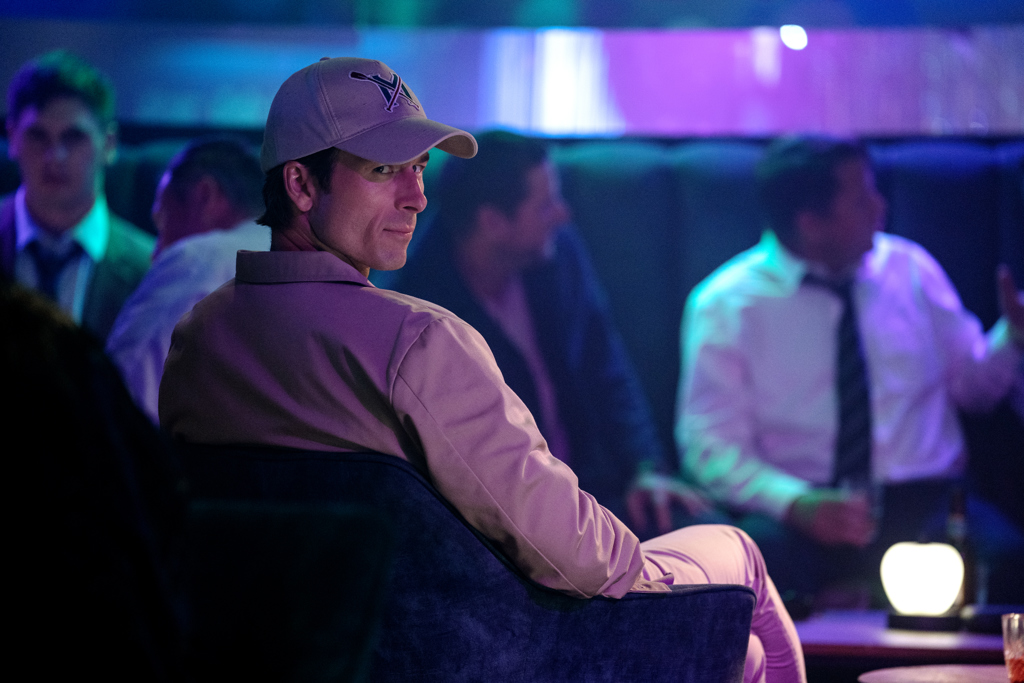 Glenn Powell as Beckett sits in a plush chair in a high-end bar environment in a scene from How to Make a Killing