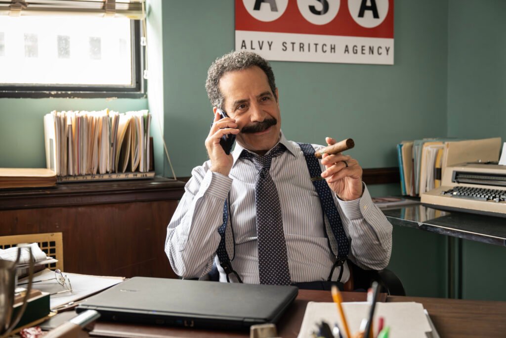 Tony Shalhoub as Alvy Stritch with a phone in his left hand held to his ear and a cigar in his right hand, sat behind a desk in American Classic