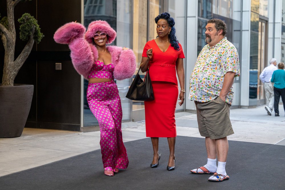 Precious Way as Brina in a pink ensemble, Erika Alexander in a red dress, and Bobby Moynihan in a multi-coloured shirt and khaki shorts stood next to each other in front of glass windows and a plant.