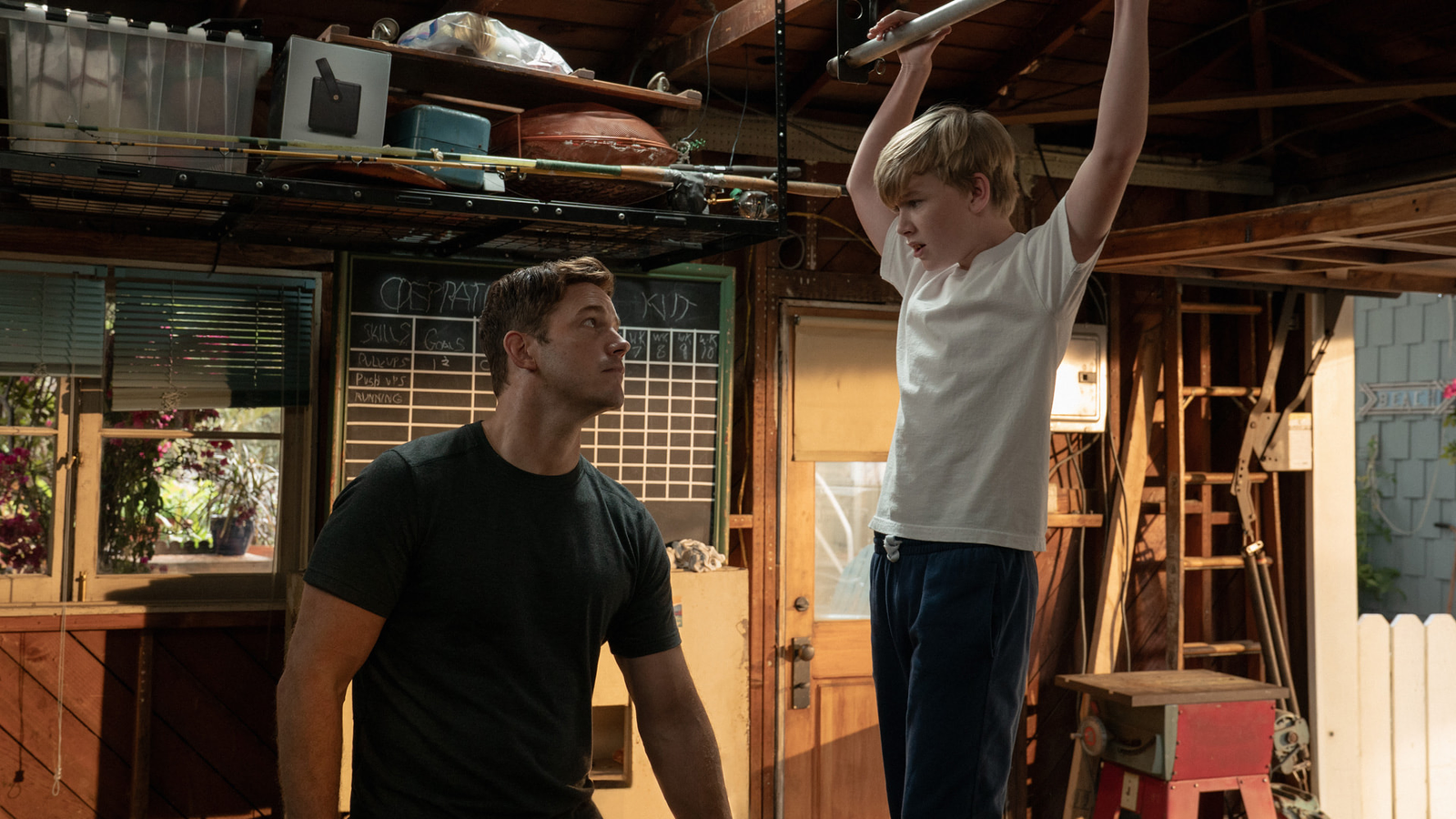 From Way of the Warrior Kid. In a garage, Chris Pratt wears a black t-shirt and looks up at Jude Hill, who is hanging from the rafters.