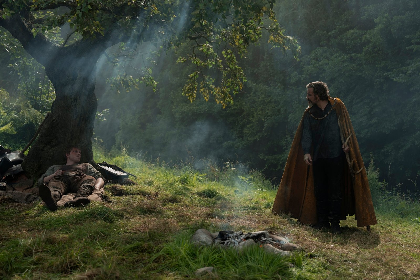 Peter Claffey as Ser Duncan the Tall lays against a tree by a dying fire while Daniel Ings as Lyonel Baratheon looks on in A Knight of the Seven Kingdoms.