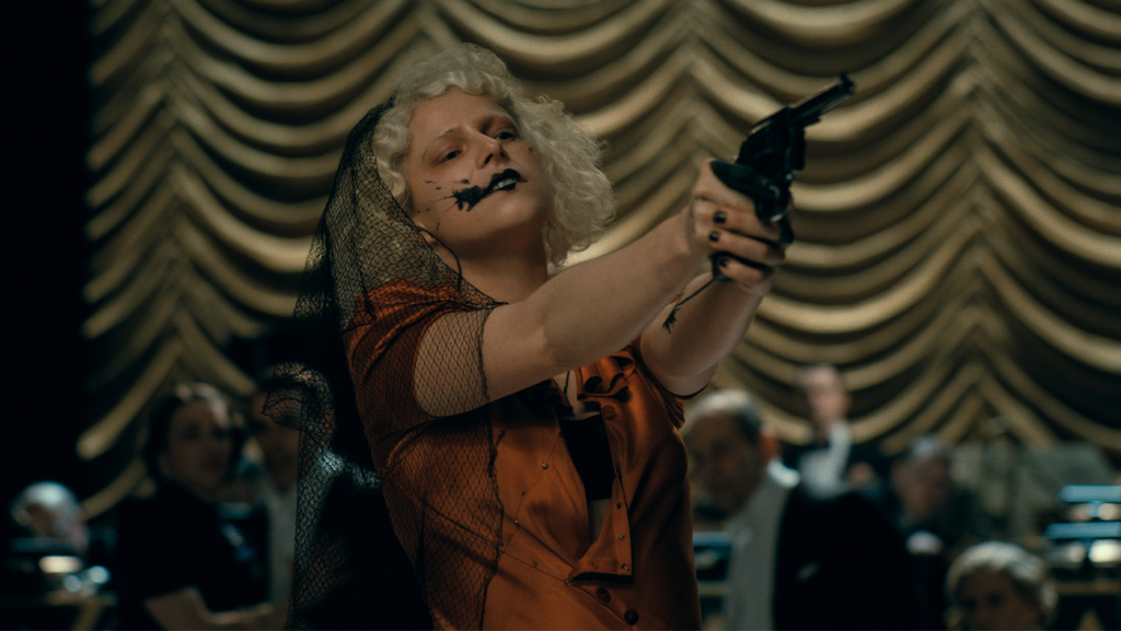 Jessie Buckley as Ida/Penney in The Bride! holding a gun in a crowded room.